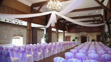 Un salón de banquetes con filas de sillas cubiertas de tela blanca y morada, un candelabro y una pared de piedra.