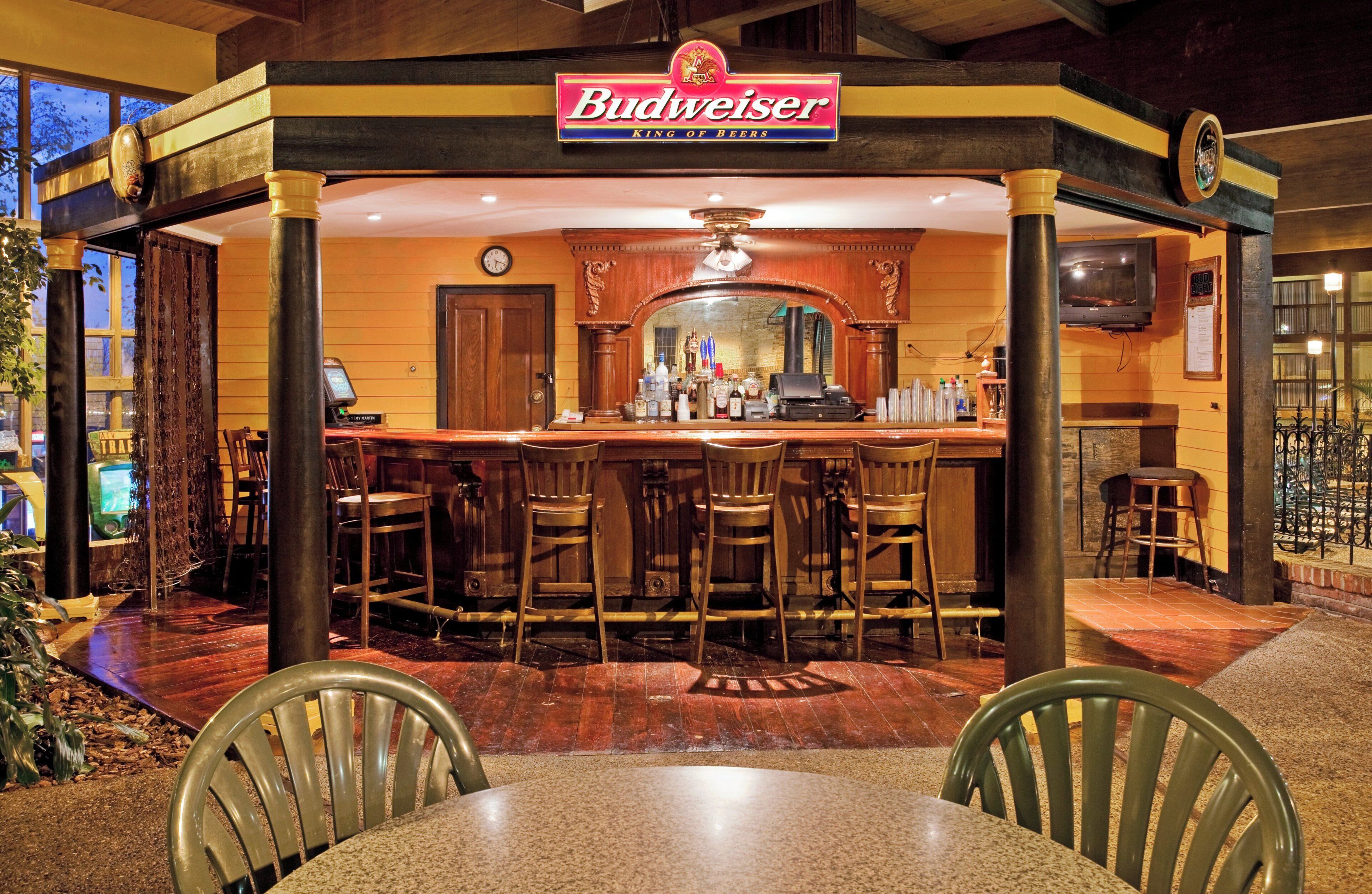 A bar area with a Budweiser sign, wooden bar counter, stools, and a clock on the wall.
