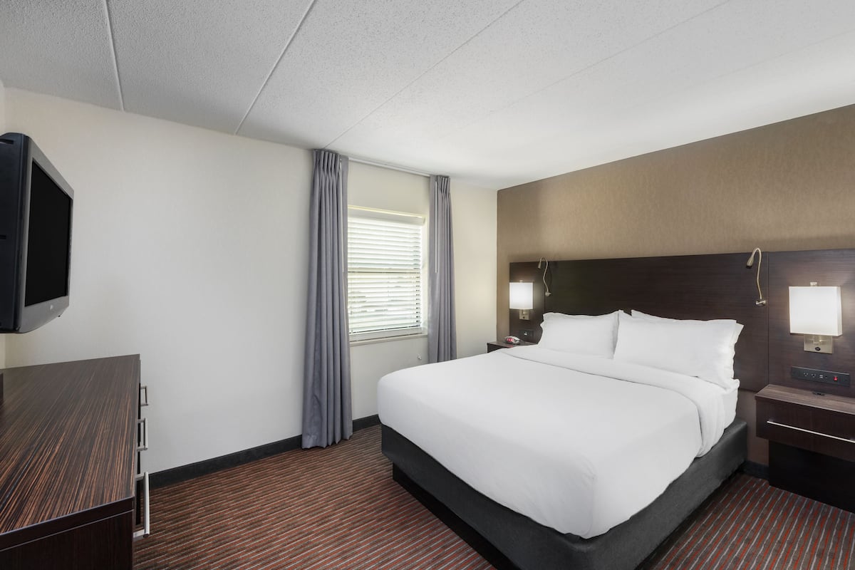 A hotel room with a large bed, a TV mounted on the wall, a desk with a chair, and a window with curtains.