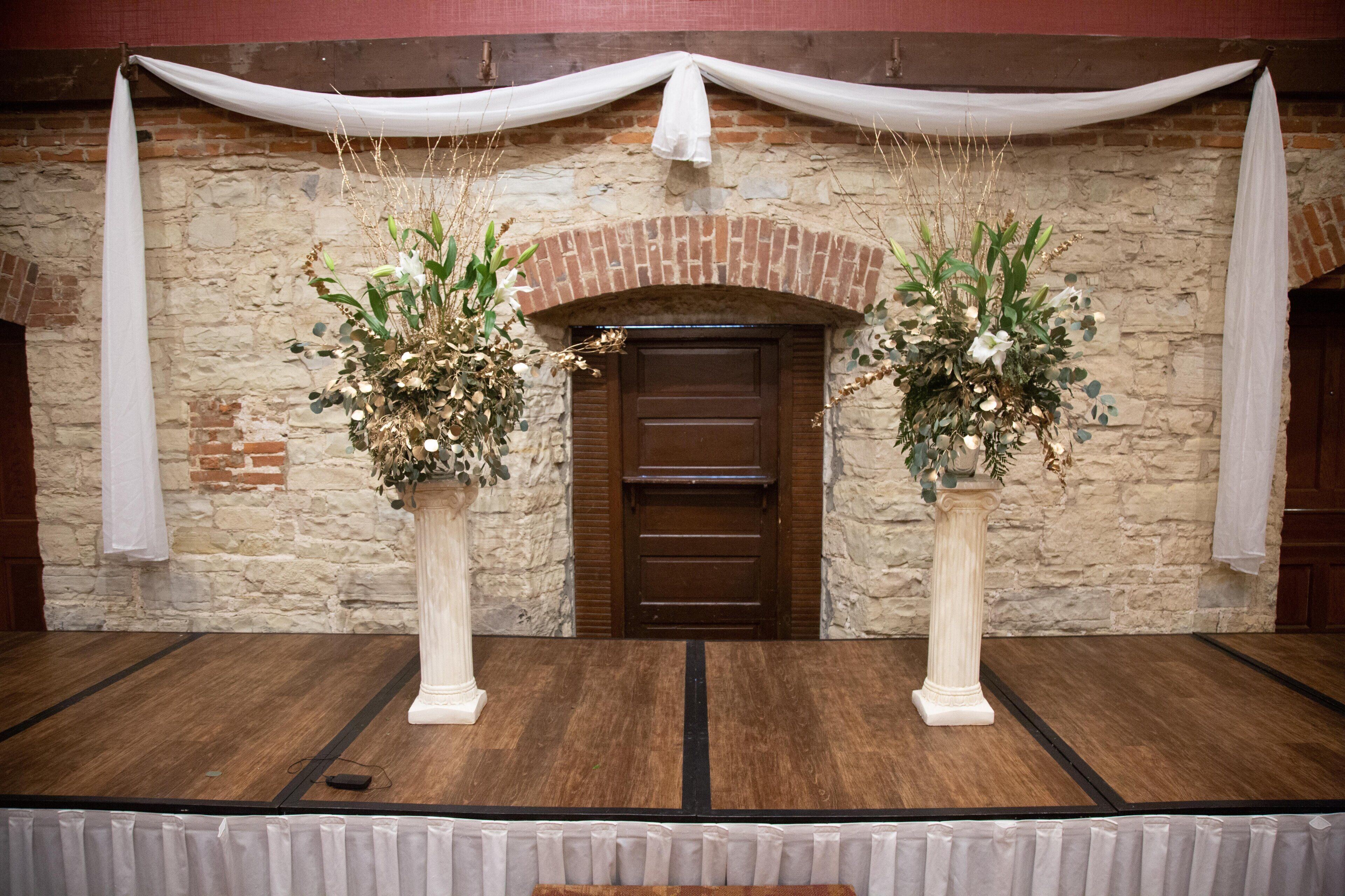 Une scène avec un décor composé d’un mur en briques, de deux colonnes blanches ornées de compositions florales et d’une porte marron.