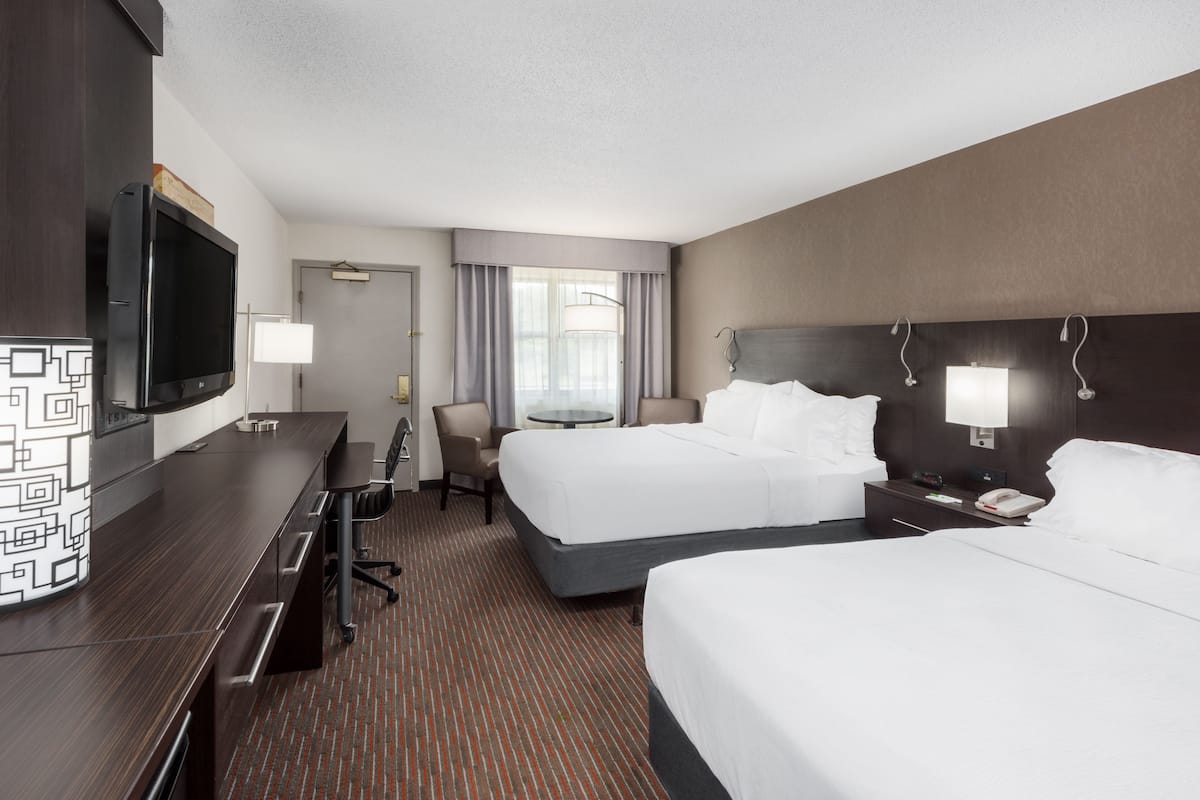 A hotel room with two beds, a desk with a flat-screen TV, a chair, a small table, and a lamp.