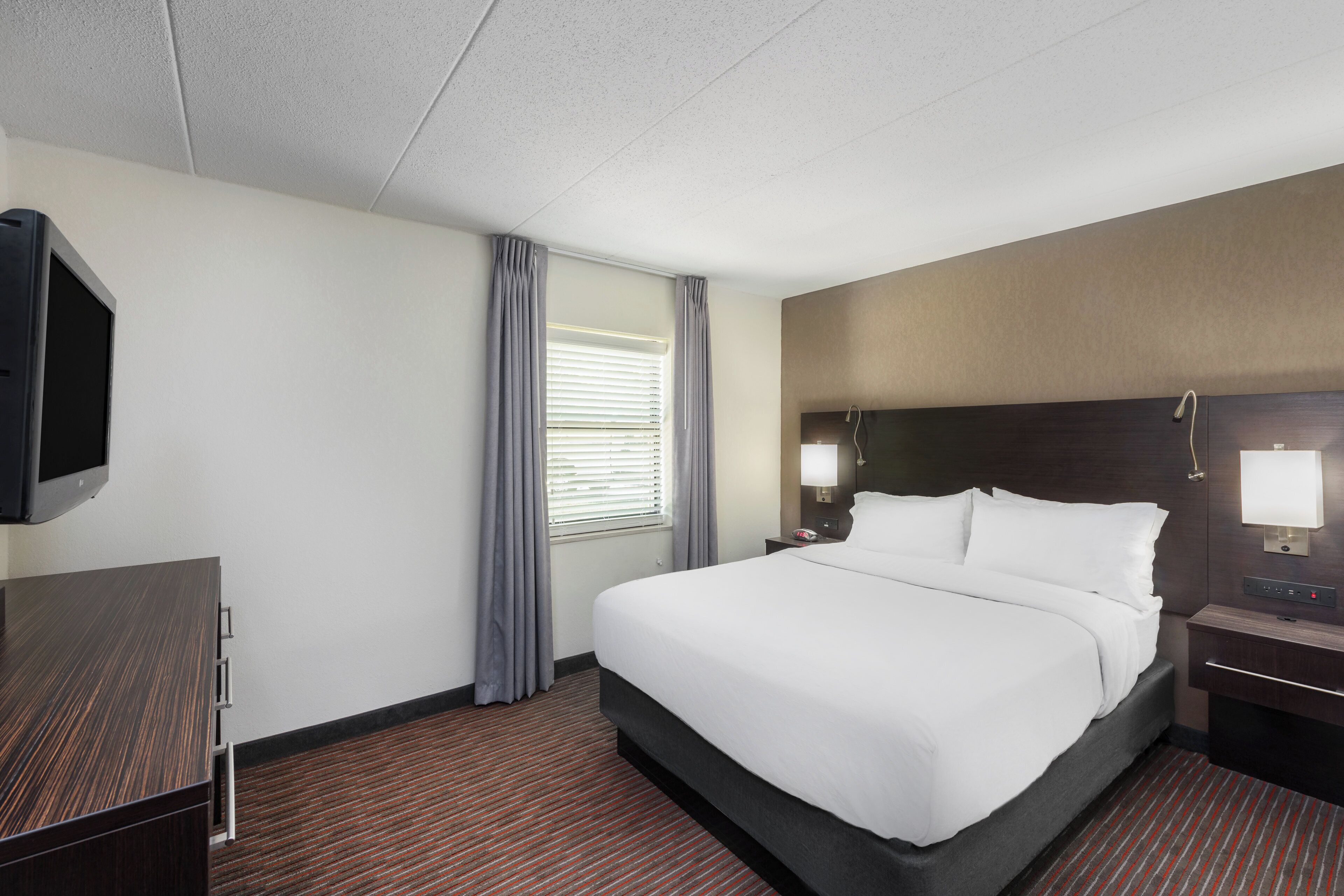 A hotel room with a bed, a TV, a nightstand, a lamp, and a window with curtains.