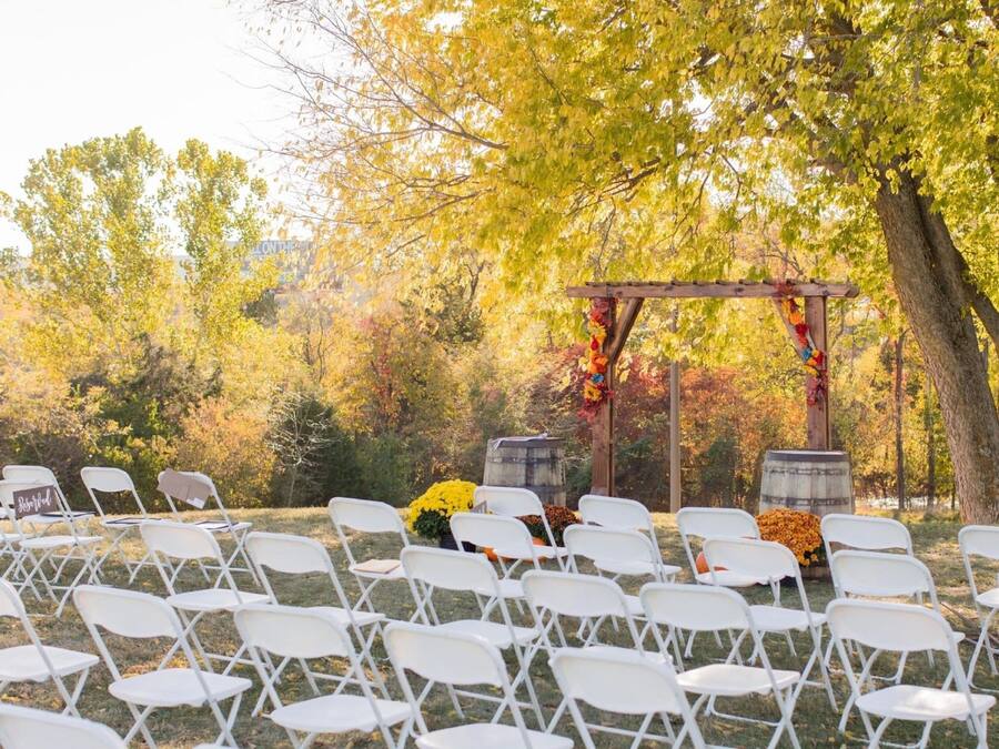 A wedding ceremony setup with white chairs arranged in rows, a wooden arch adorned with flowers, and a backdrop of autumn trees.