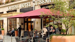Uteservering med ett rött parasoll, en skylt med texten ”Kastens Hotel Luisenhof” och utsikt över byggnadens entré.