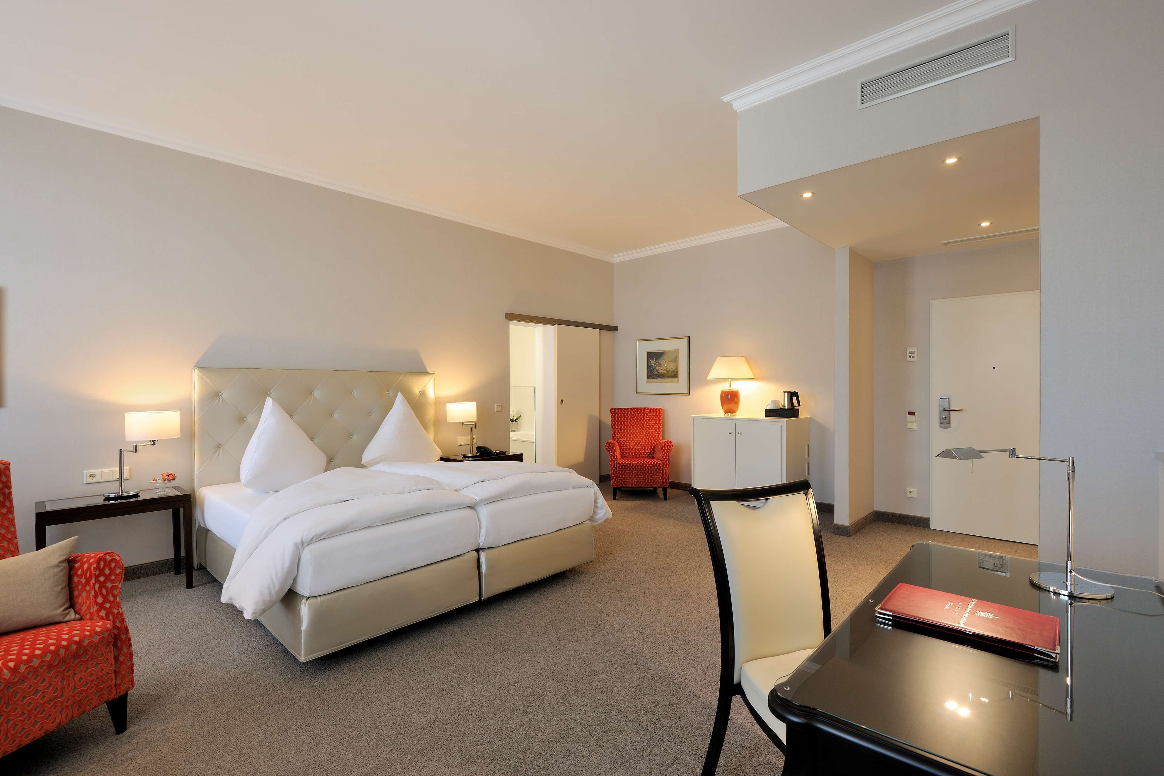 A hotel room with a bed, a desk, a chair, a red armchair, and a lamp.