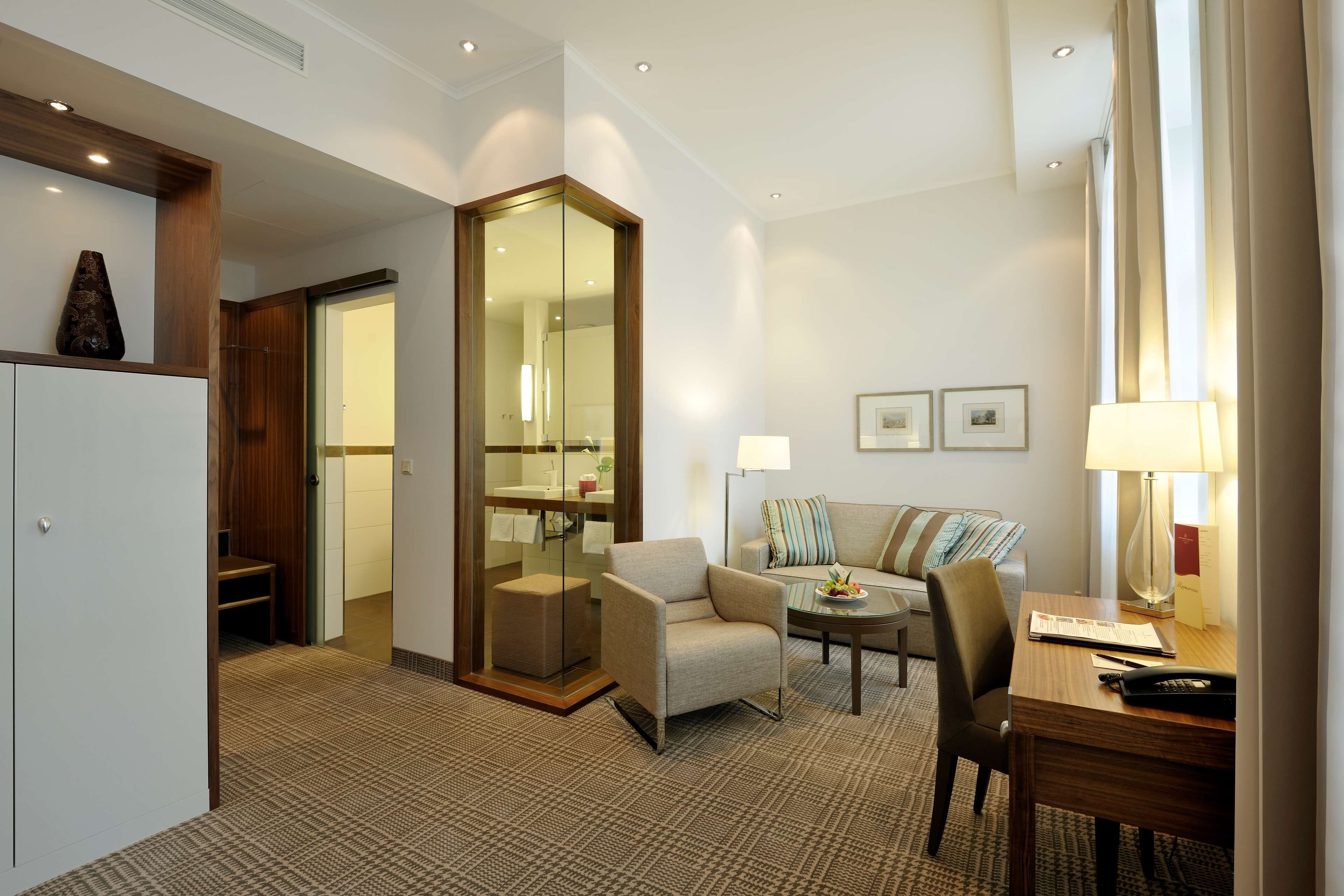 Ein Hotelzimmer mit Sofa, Ohrensessel, Esstisch und einem durch eine Glasscheibe abgetrennten Badezimmer.