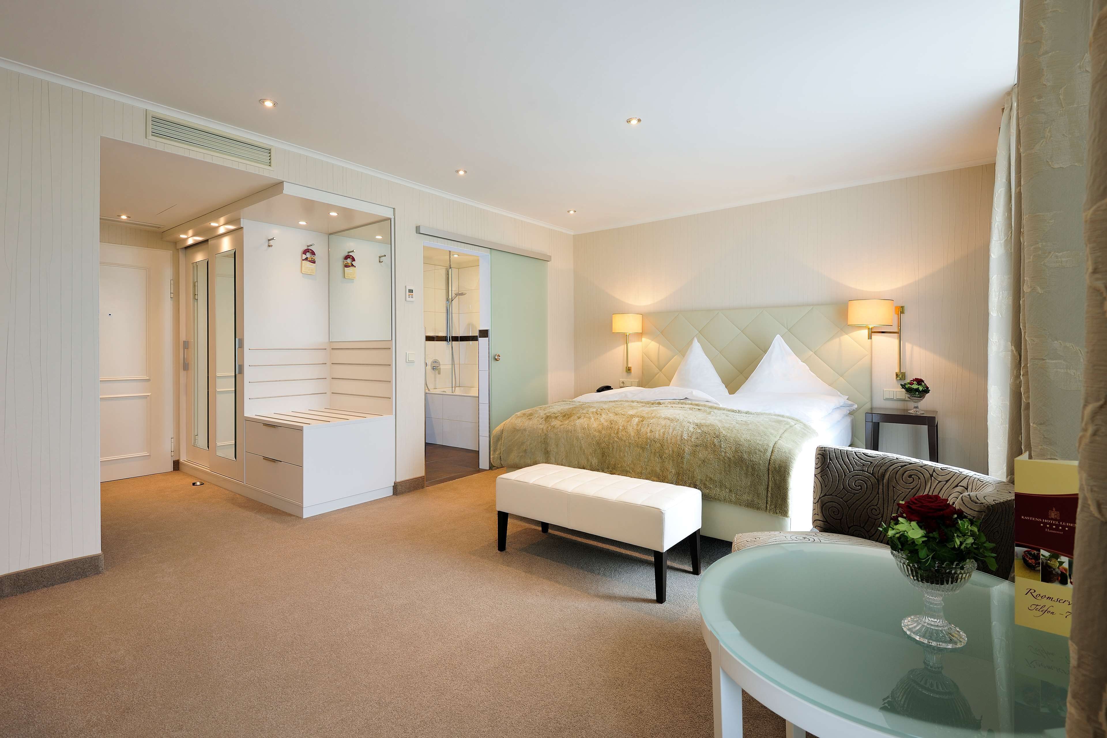 A hotel room with a bed, a bench, a glass table with a vase, a nightstand, and a mirror.
