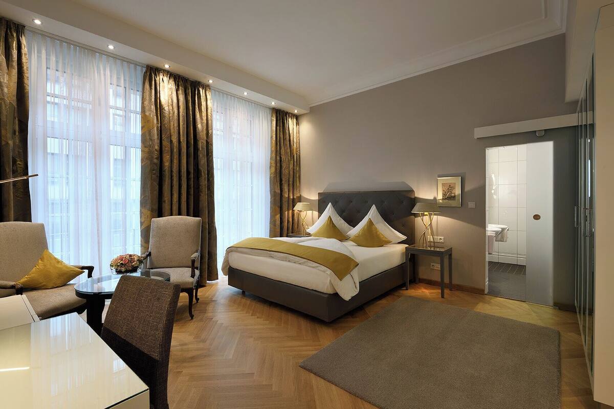 Een hotelkamer met een bed, twee fauteuils, een kleine tafel met een vaas en een nachtkastje met een lamp.