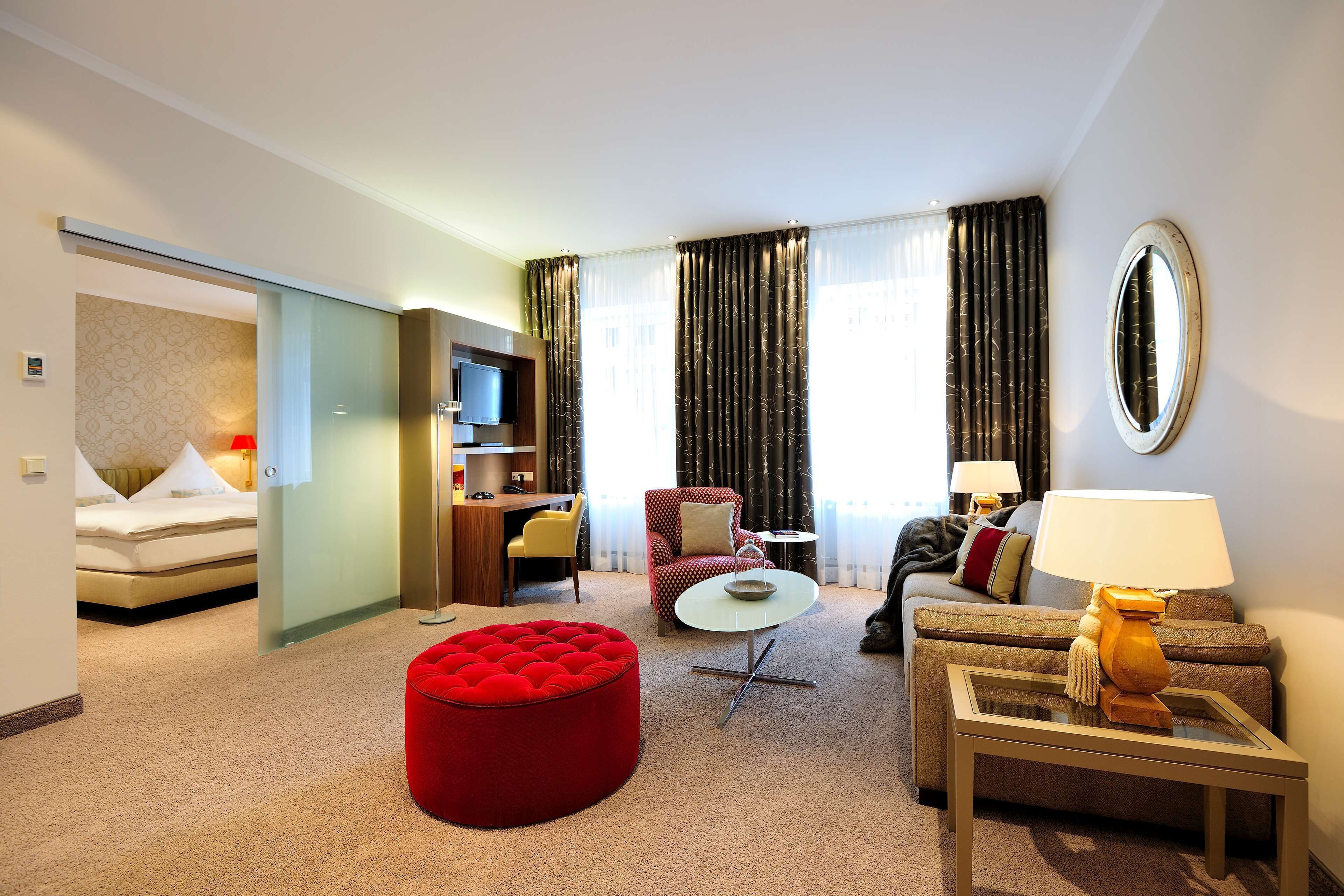 Une chambre d’hôtel avec un lit, un pouf rouge, un canapé, une table basse, une télévision et un bureau.