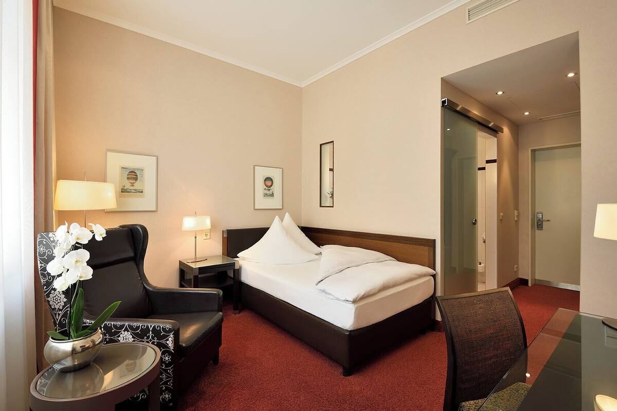 Een hotelkamer met een bed, een bureau, een stoel, een kleine tafel met een bloemenvaas, en een deur die naar een andere kamer leidt.