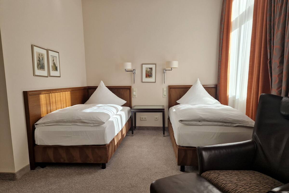 Een hotelkamer met twee eenpersoonsbedden, een klein tafeltje, twee wandlampen en een leren fauteuil.