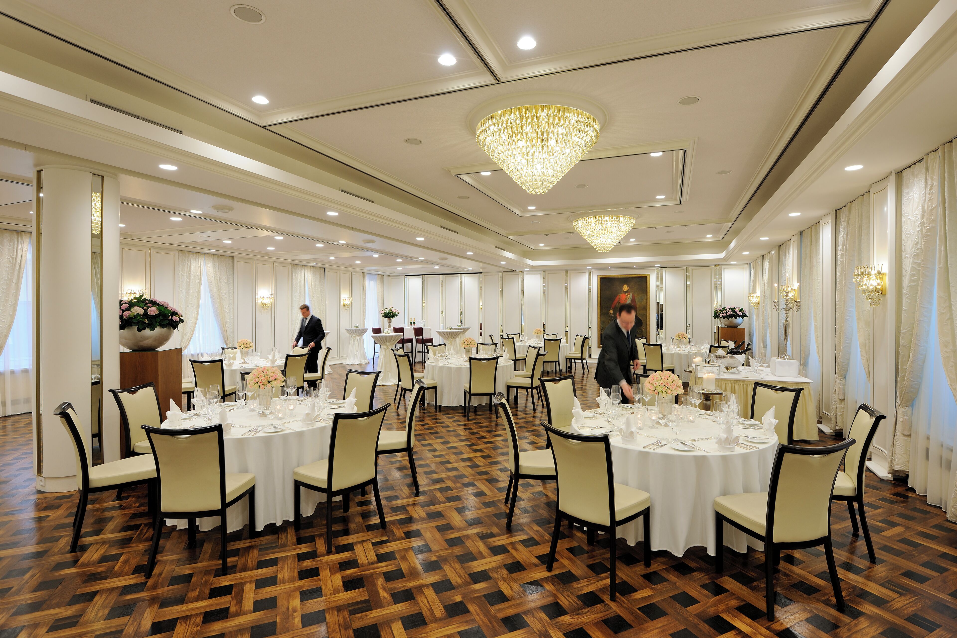 Un salón de banquetes con mesas redondas dispuestas para comer, candelabros y un piso de madera pulida.