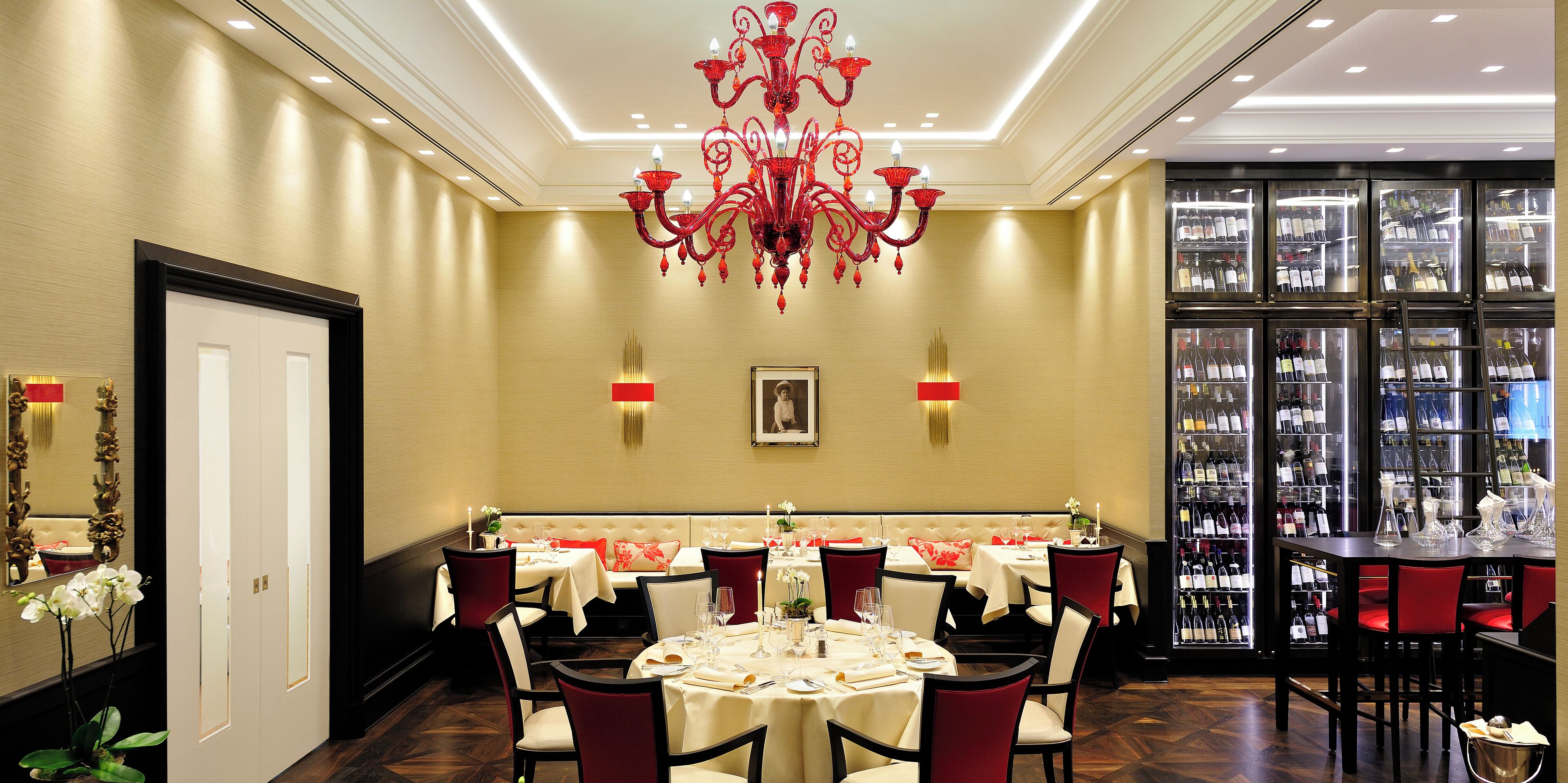 A hotel dining area with round tables, red upholstered chairs, a central chandelier, and a wine display case in the background.
