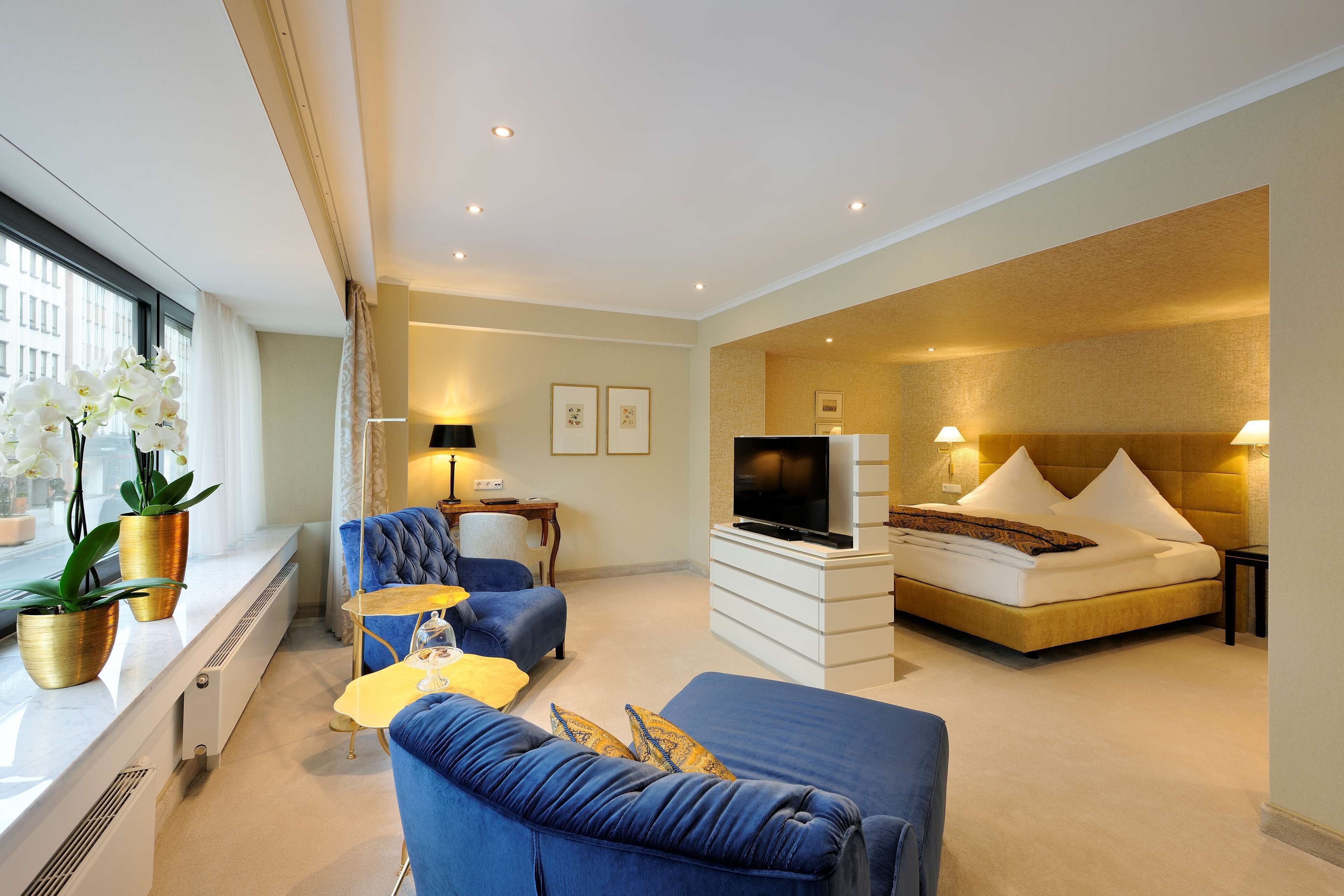 Habitación de hotel con cama, televisor, sofá, mesa de centro y ventana con plantas.