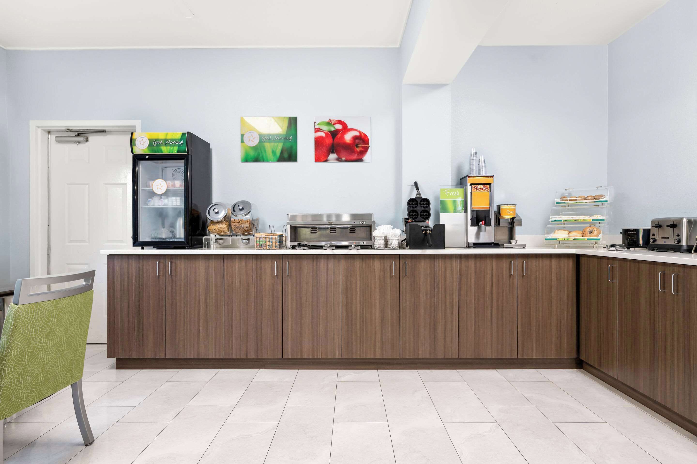 Een moderne keuken met een inbouwkoelkast, magnetron en koffieapparaat. Twee wanddecoraties: één met appels en één met een groene achtergrond.
