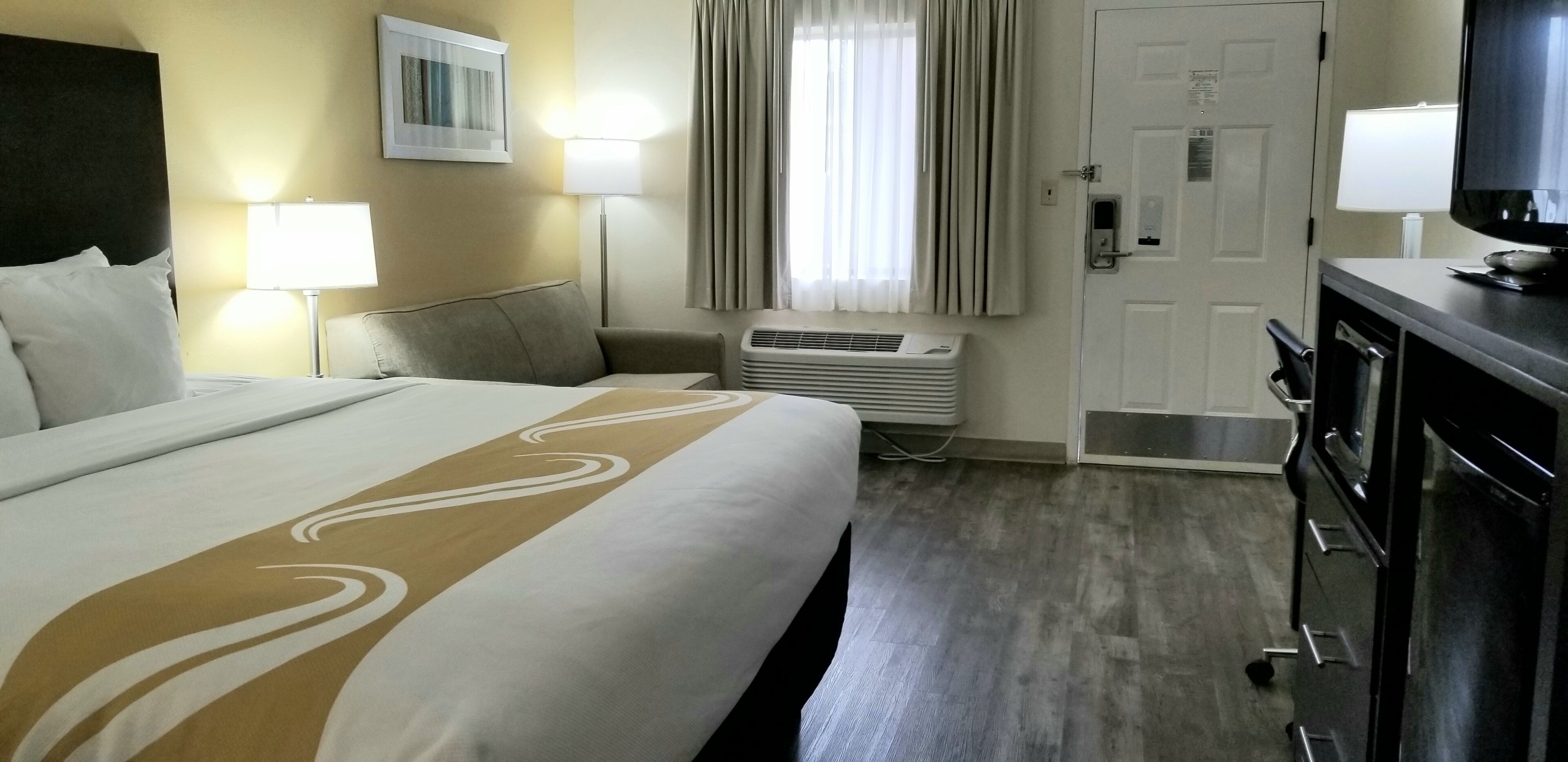 Una habitación de hotel con una cama grande, un escritorio con televisor, una lámpara de escritorio, una ventana con cortinas y un equipo de aire acondicionado.
