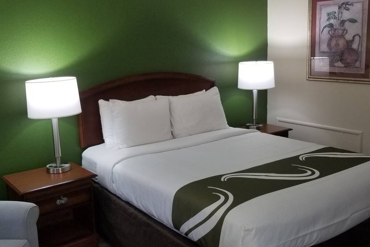 A neatly made bed with white linens and a green accent, two bedside lamps, a wooden nightstand, and a framed artwork on the wall.