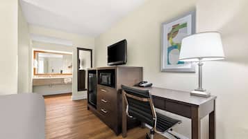 A hotel room with a wooden floor, a desk with a computer, a chair, a TV, a lamp, and a bathroom visible in the background.