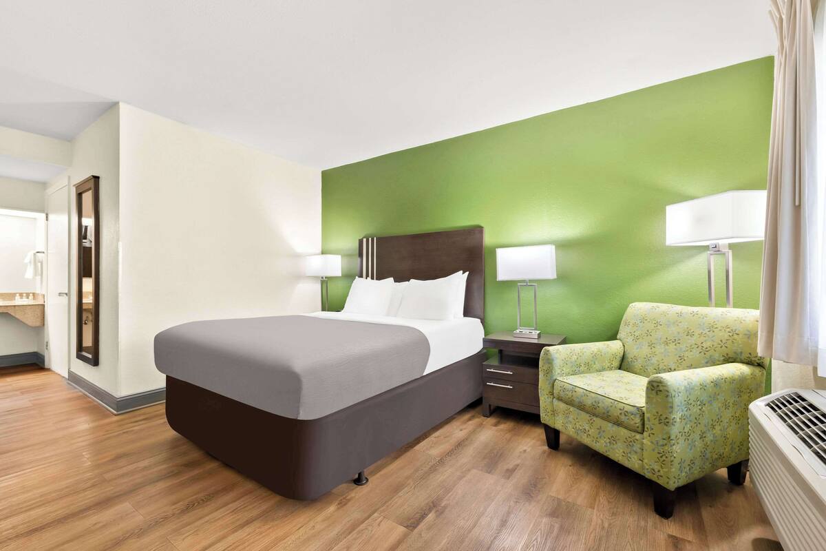 A hotel room with a large bed, a nightstand, a chair, and a green accent wall.