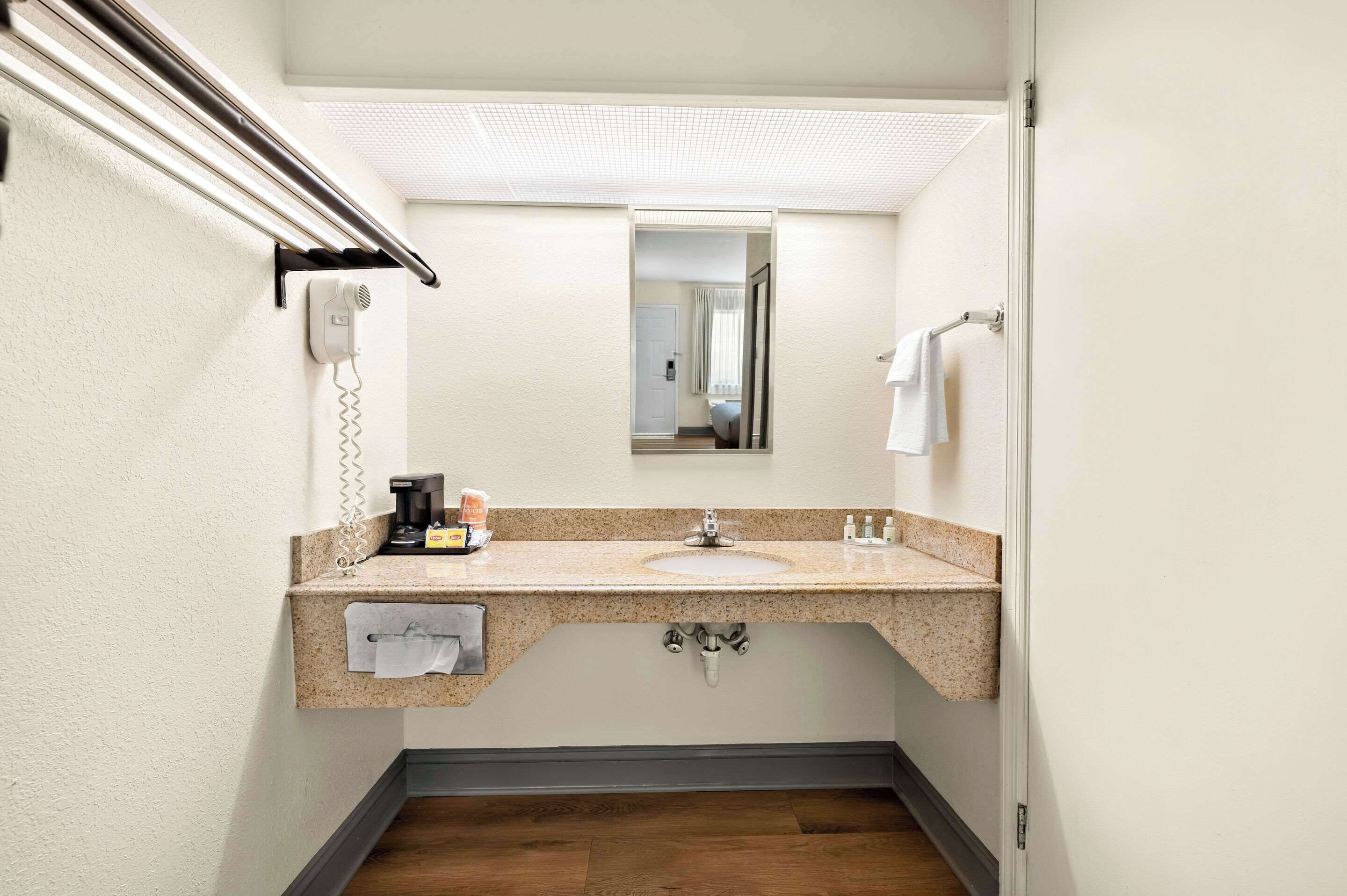 A compact bathroom with a granite countertop sink, a mirror, a towel rack, a hairdryer, and a coffee maker.