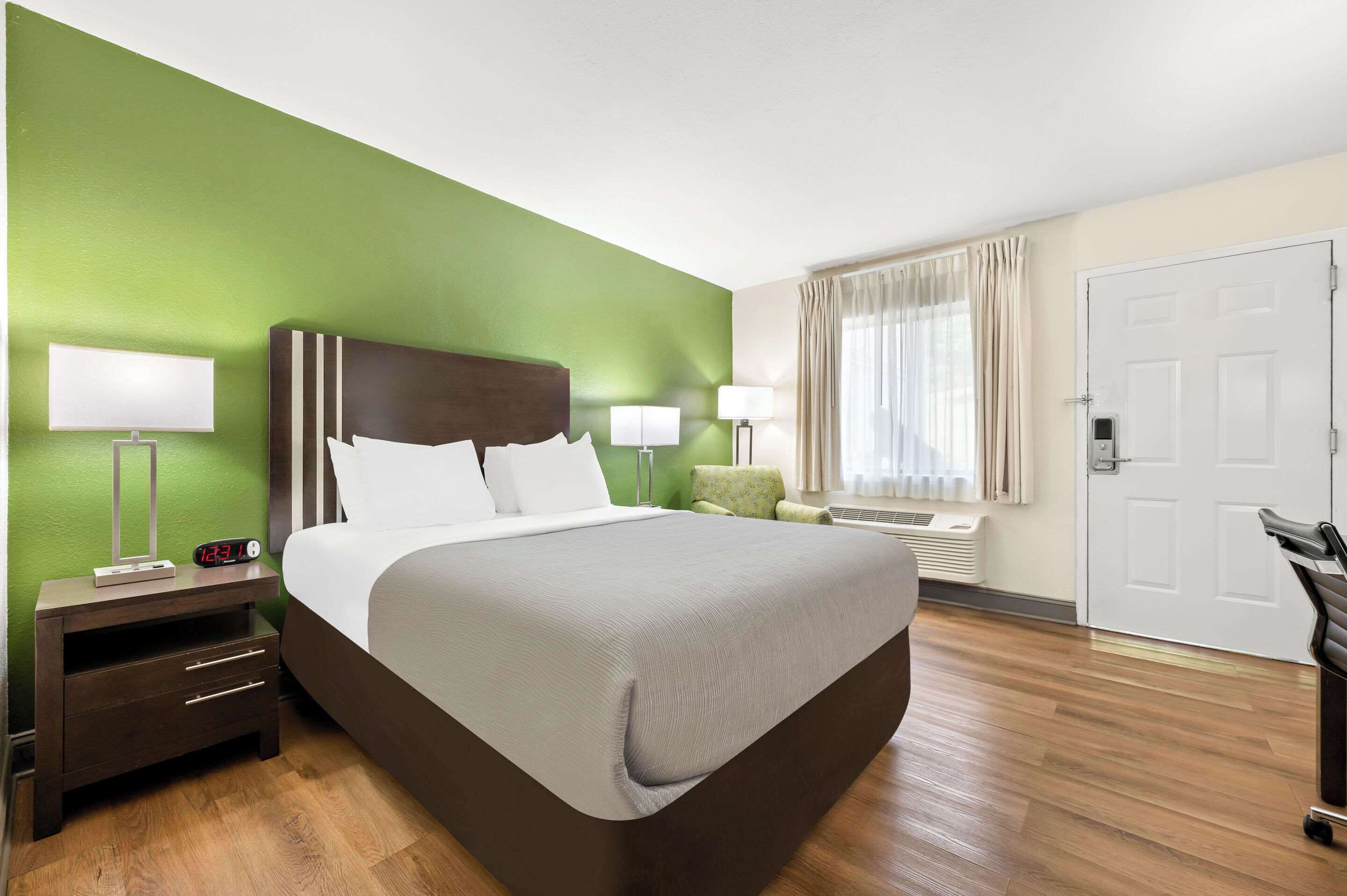 Quarto de hotel com uma cama grande, luminárias de cabeceira, mesa de cabeceira, parede verde e uma janela com cortinas, além de uma porta.