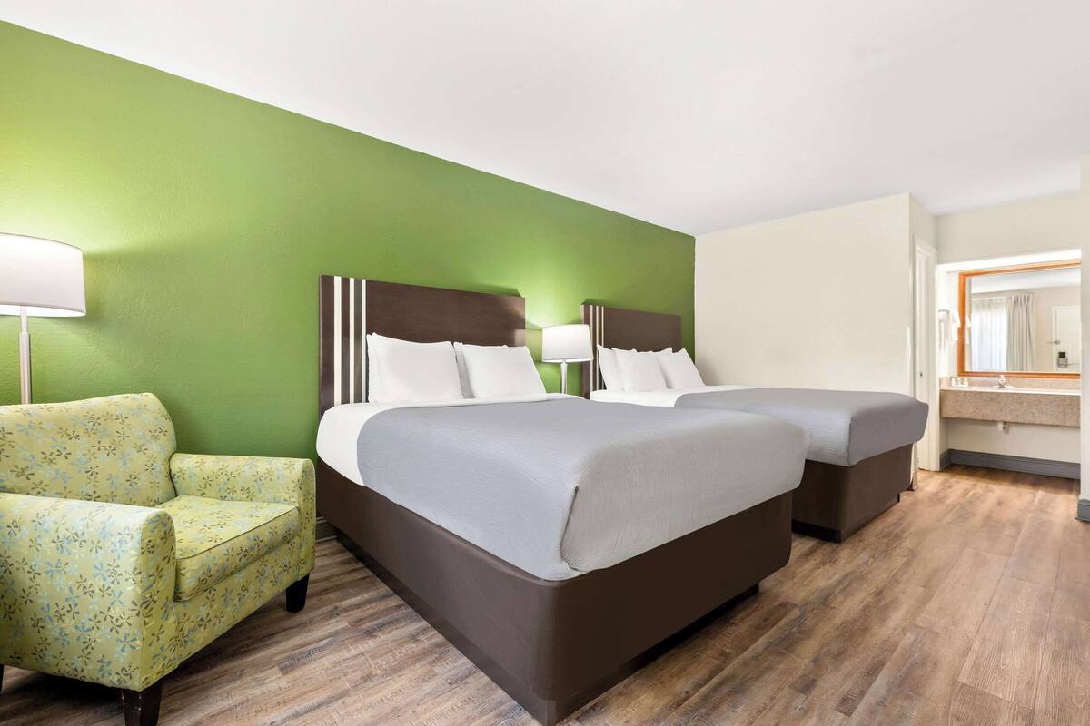 A hotel room with two beds, a green wall, a wooden floor, a chair, and a bathroom with a mirror and sink.