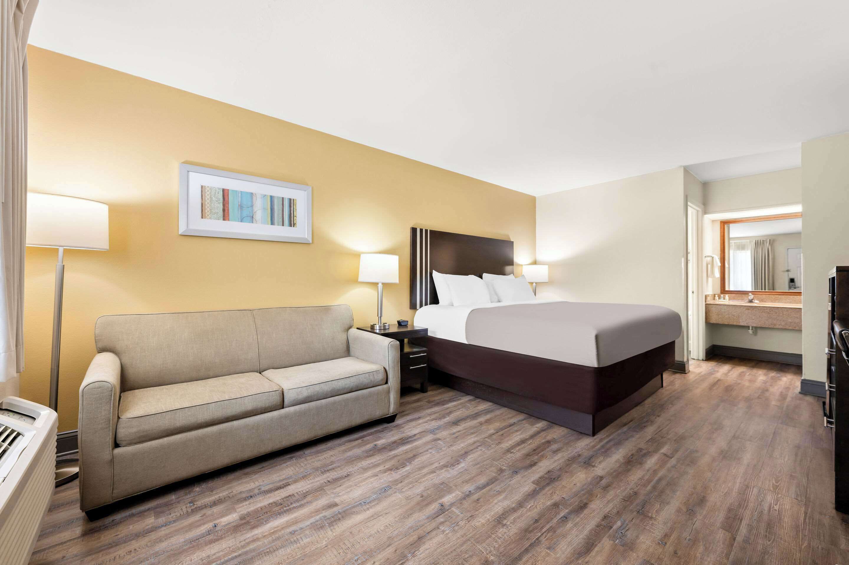 A hotel room with a bed, a sofa, a nightstand, a lamp, a painting, and a bathroom visible through an open door.