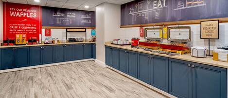 A breakfast buffet area with a variety of food options, including waffles, and a sign indicating breakfast starts here.