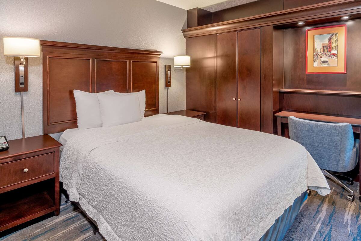 Une chambre d’hôtel avec un grand lit, une tête de lit en bois, deux tables de chevet, un bureau avec une chaise et un tableau encadré au mur.