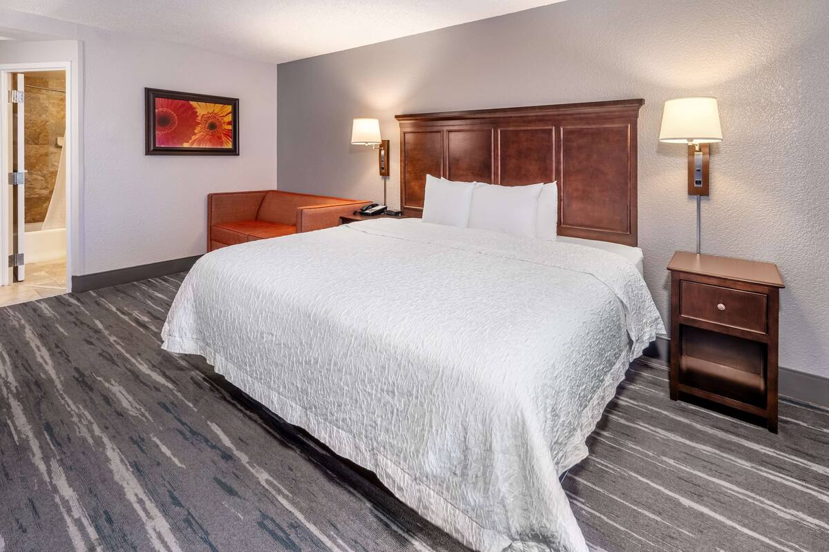 Une chambre d’hôtel avec un grand lit, une tête de lit en bois, deux lampes de chevet, une table de chevet, une petite table, un tableau au mur et une porte donnant sur une salle de bain.