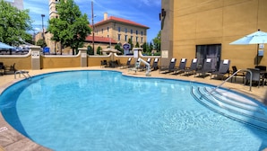 Una piscina con sillones de descanso y sombrillas, un cartel de prohibición de buceo y un edificio con tejado rojo al fondo.