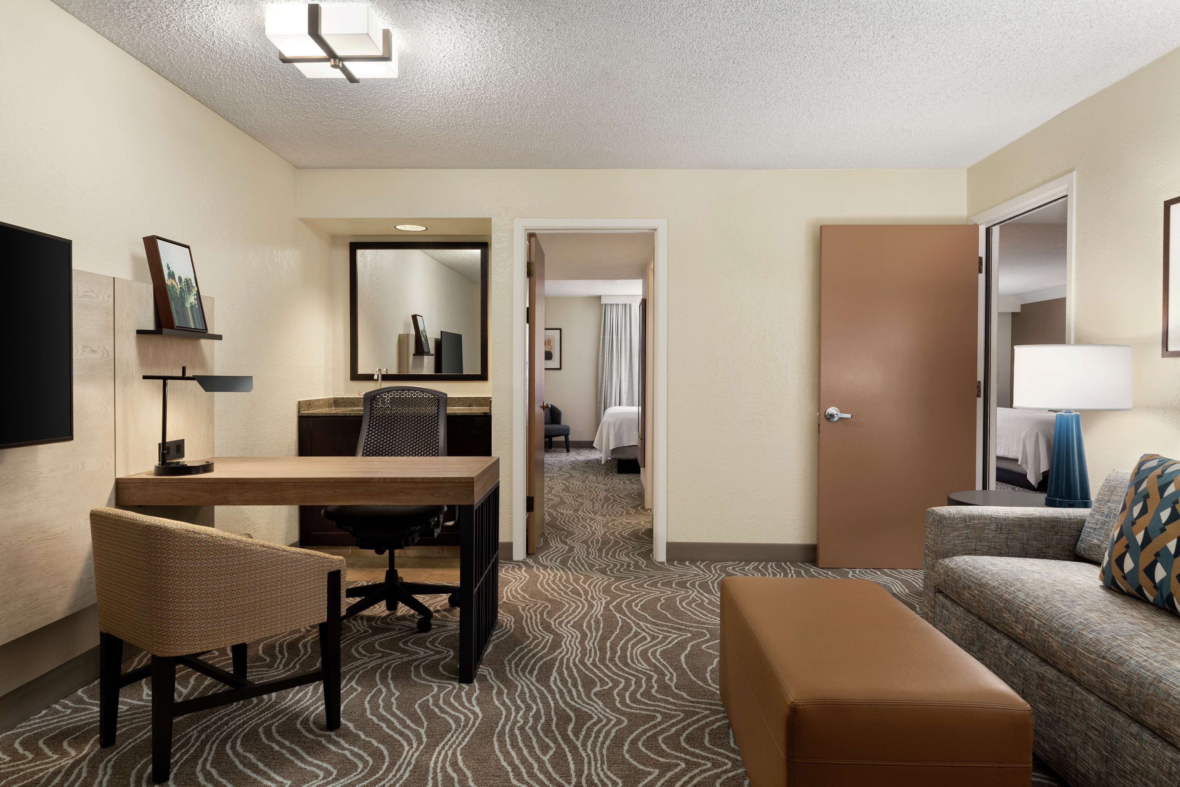 Une chambre d’hôtel équipée d’un bureau, d’une chaise, d’un canapé et d’un pouf. On y trouve également un miroir, une lampe et une porte donnant sur une autre pièce.
