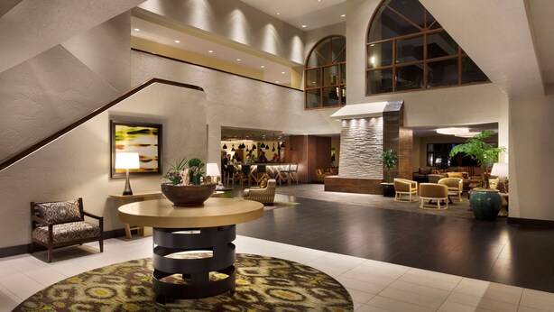 A spacious lobby with a high ceiling, a central round table, seating areas, and a staircase.