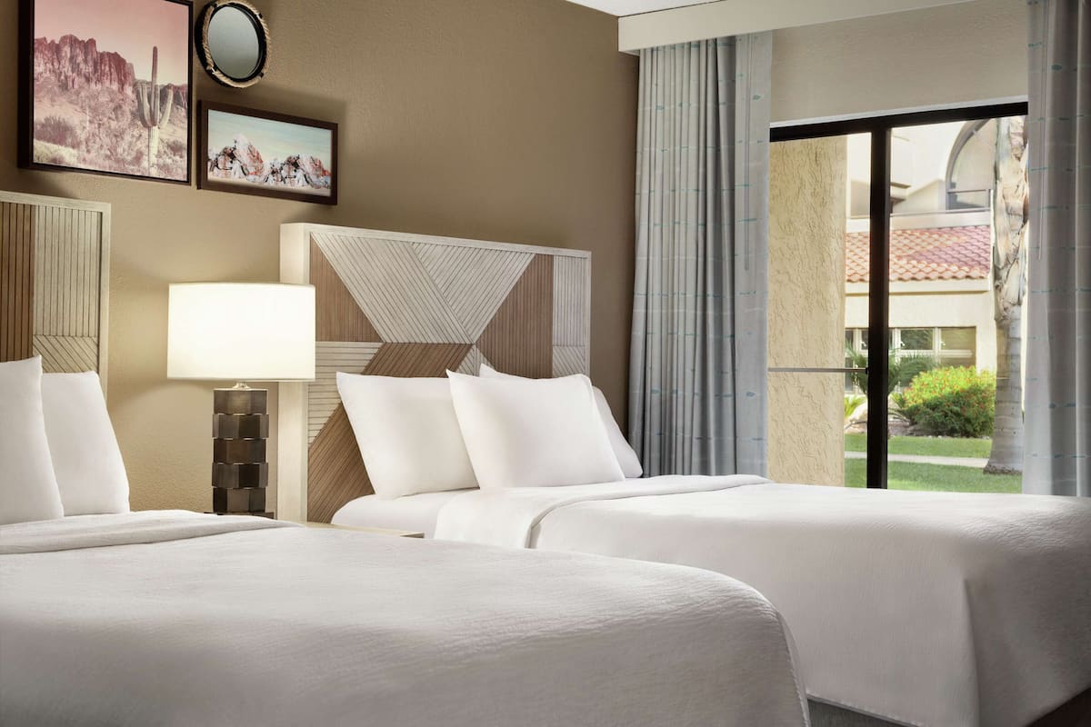A hotel room with two beds, a nightstand, a lamp, and a large window with a view of the outdoors.