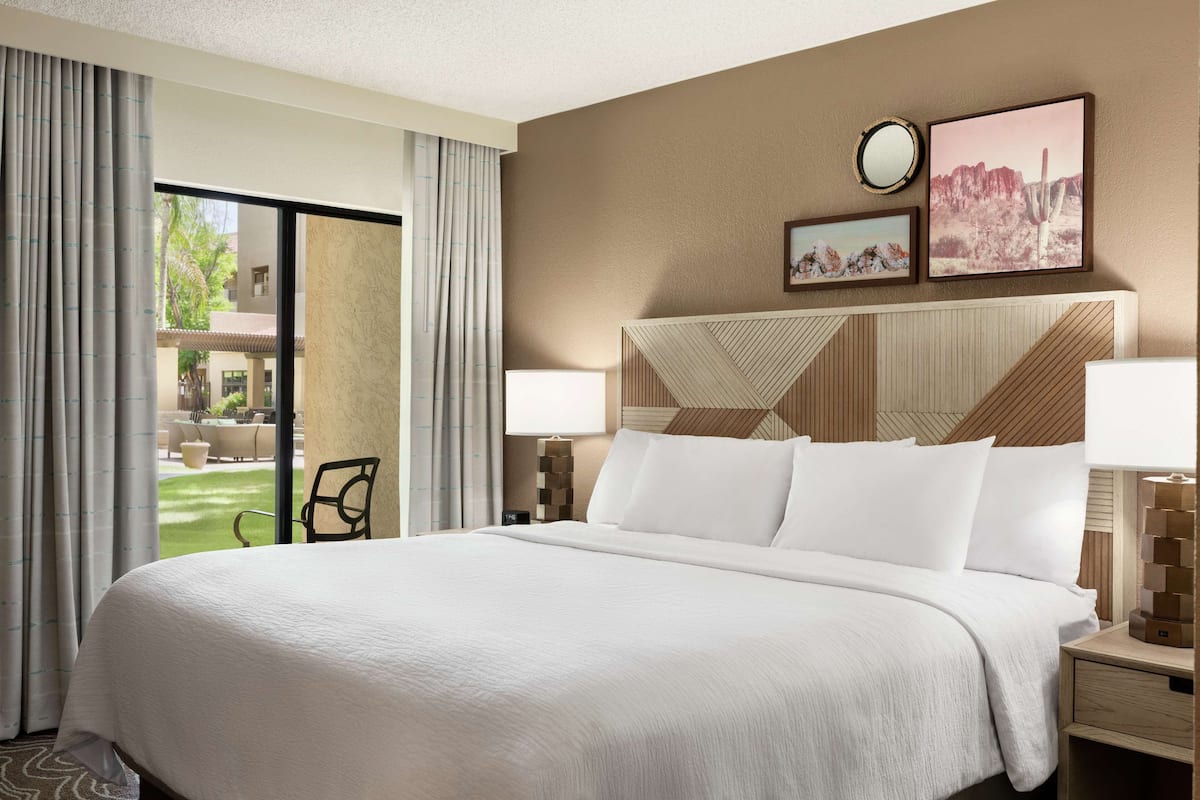 A hotel room with a large bed, two bedside tables, a view of the outdoor area, and a wall with two framed pictures.