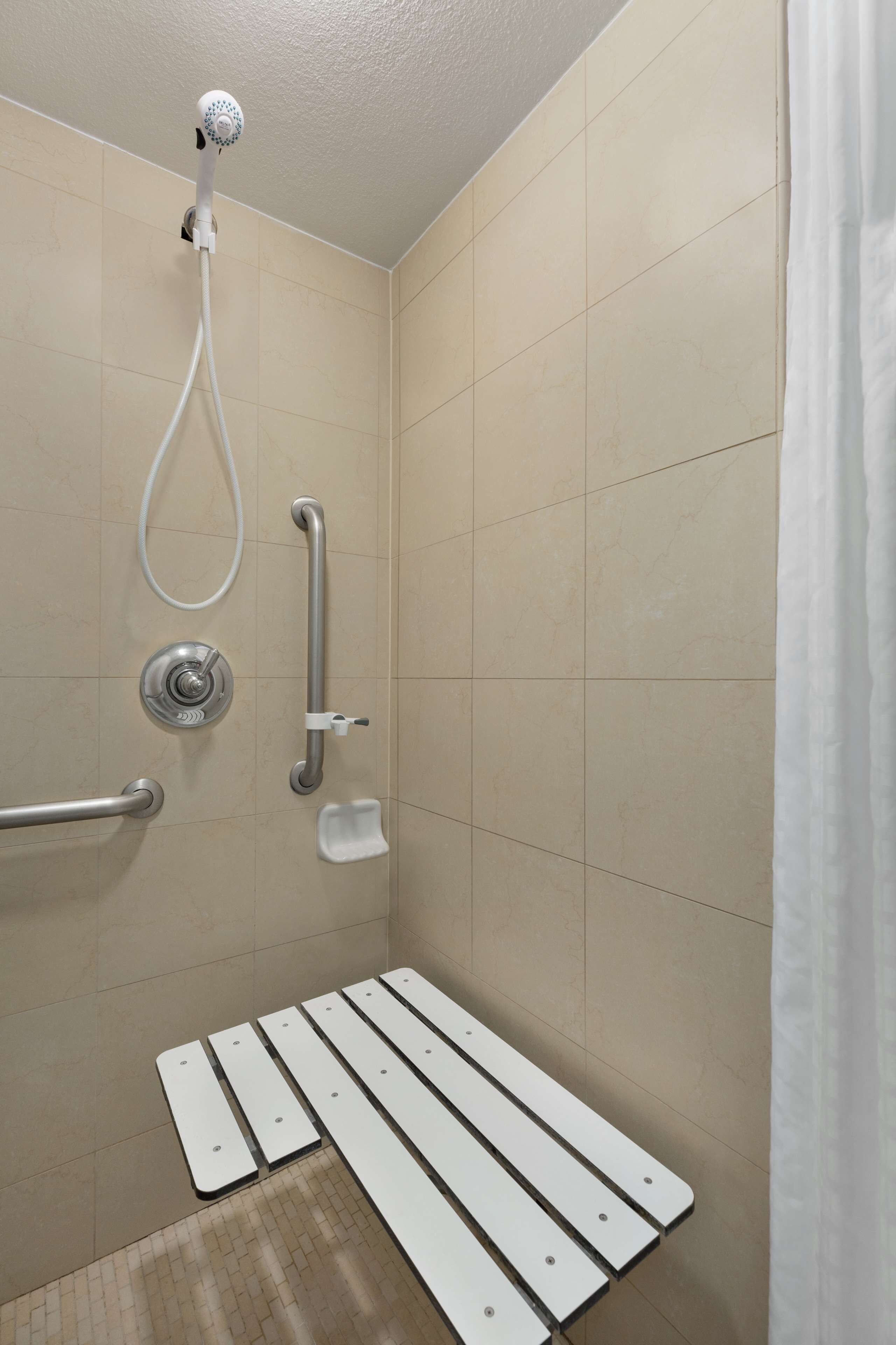 Un espacio de ducha con cabezal de ducha de mano, barras de apoyo y un asiento blanco.