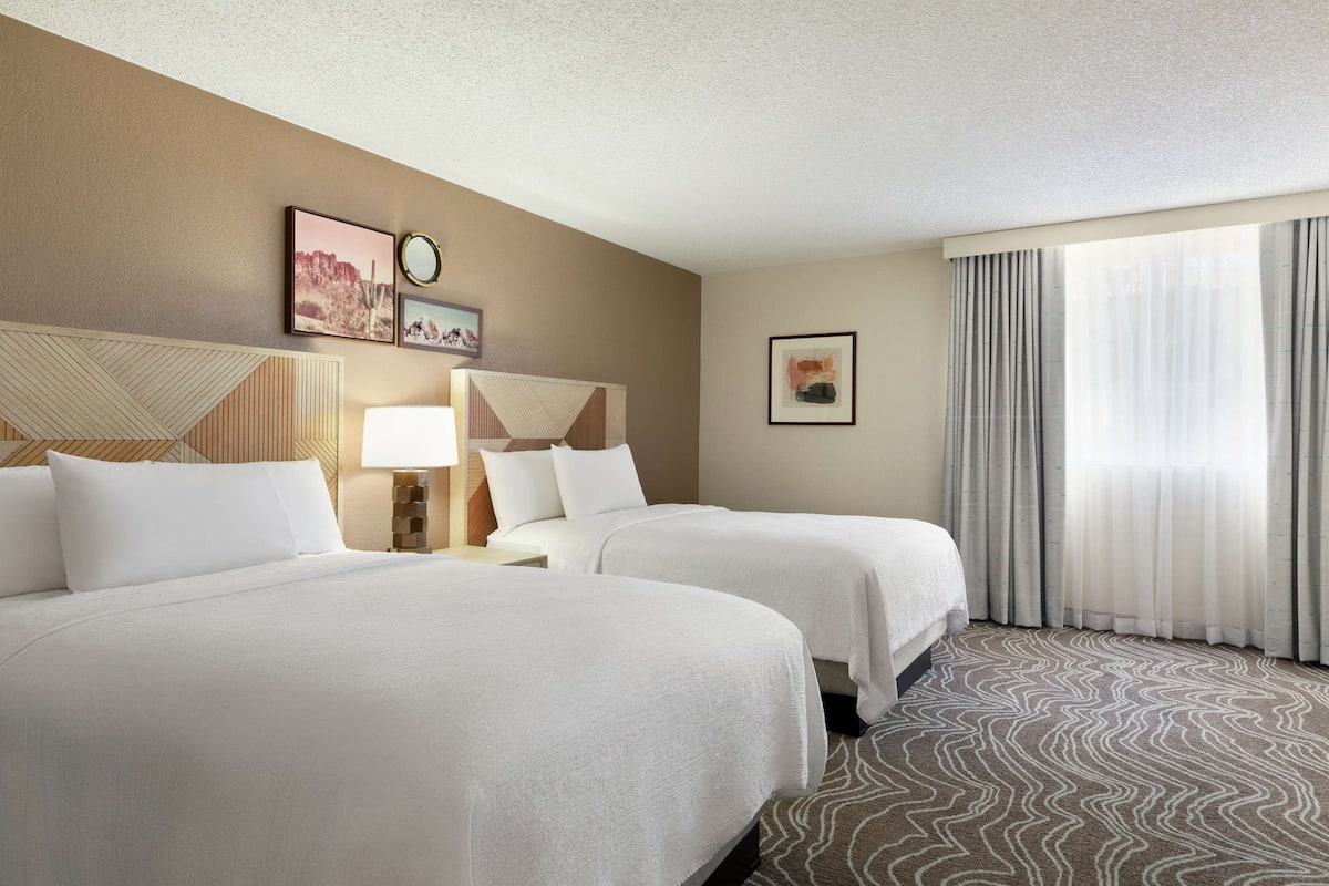 A hotel room with two beds, a large window with curtains, a wall clock, and framed artwork.