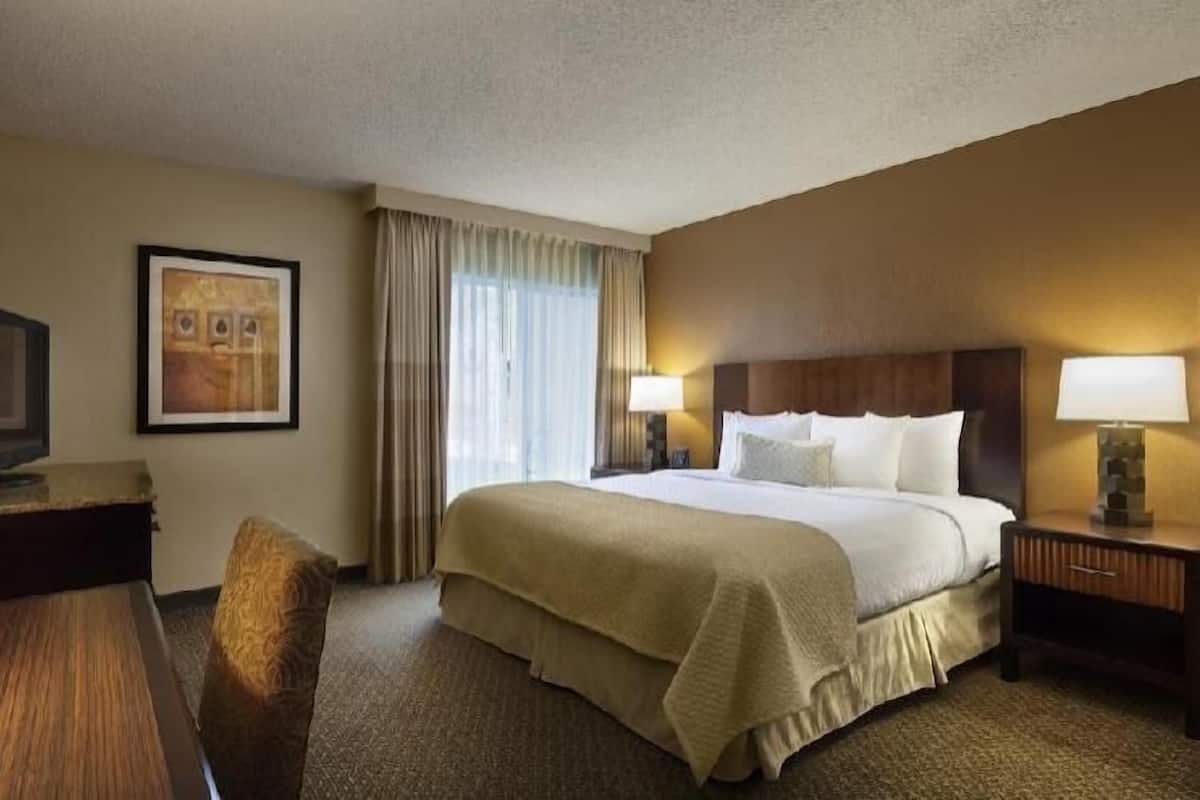 A hotel room with a bed, two bedside lamps, a desk with a television, a chair, and a framed picture on the wall.