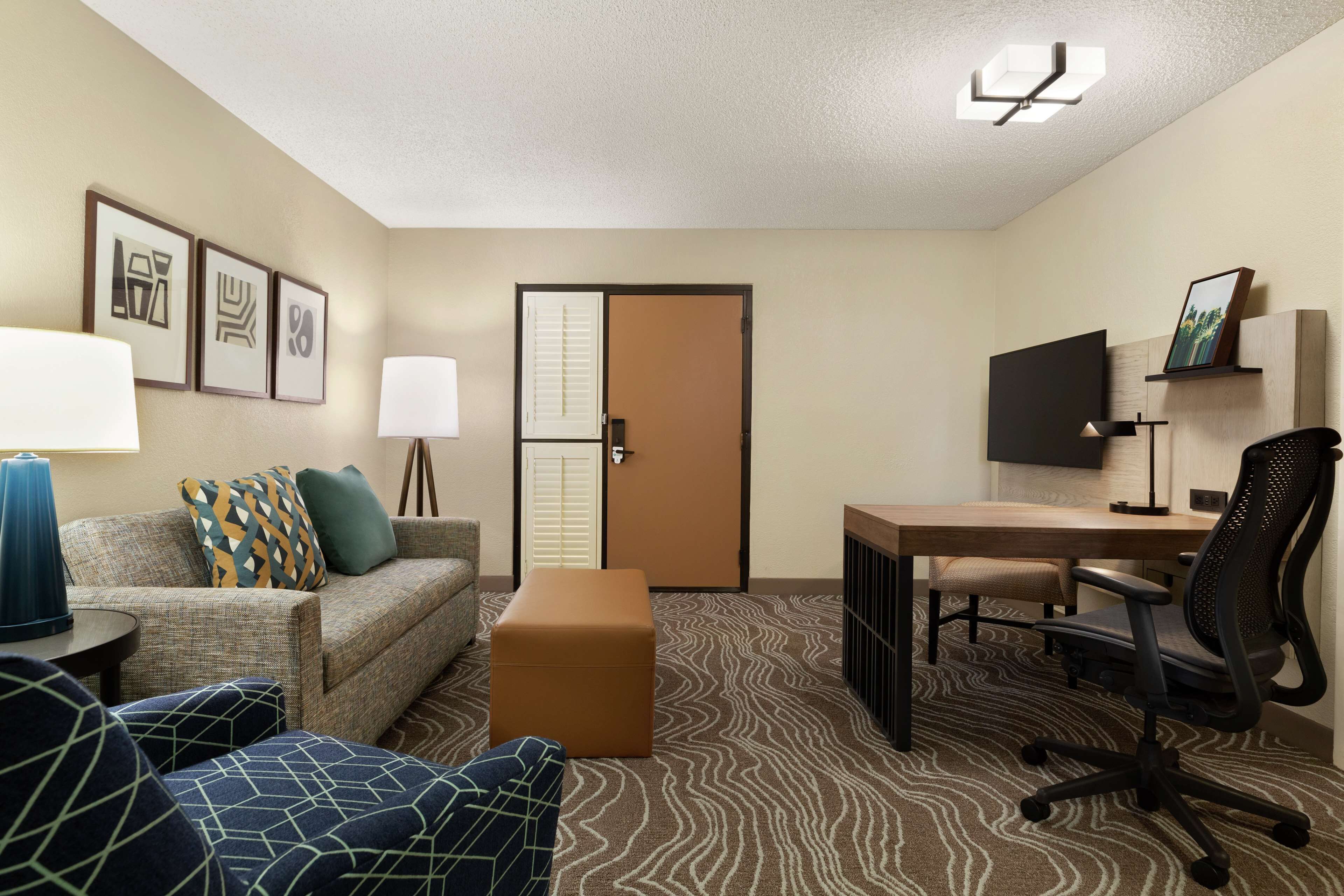A hotel room with a sofa, ottoman, desk, chair, TV, lamp, and framed pictures on the wall.