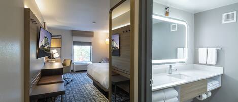 A hotel room with a bed, a desk, a chair, a large mirror, and a bathroom with a sink and towel rack.