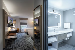 Habitación de hotel con cama, escritorio, silla, televisor y baño con lavamanos, espejo y toallero.
