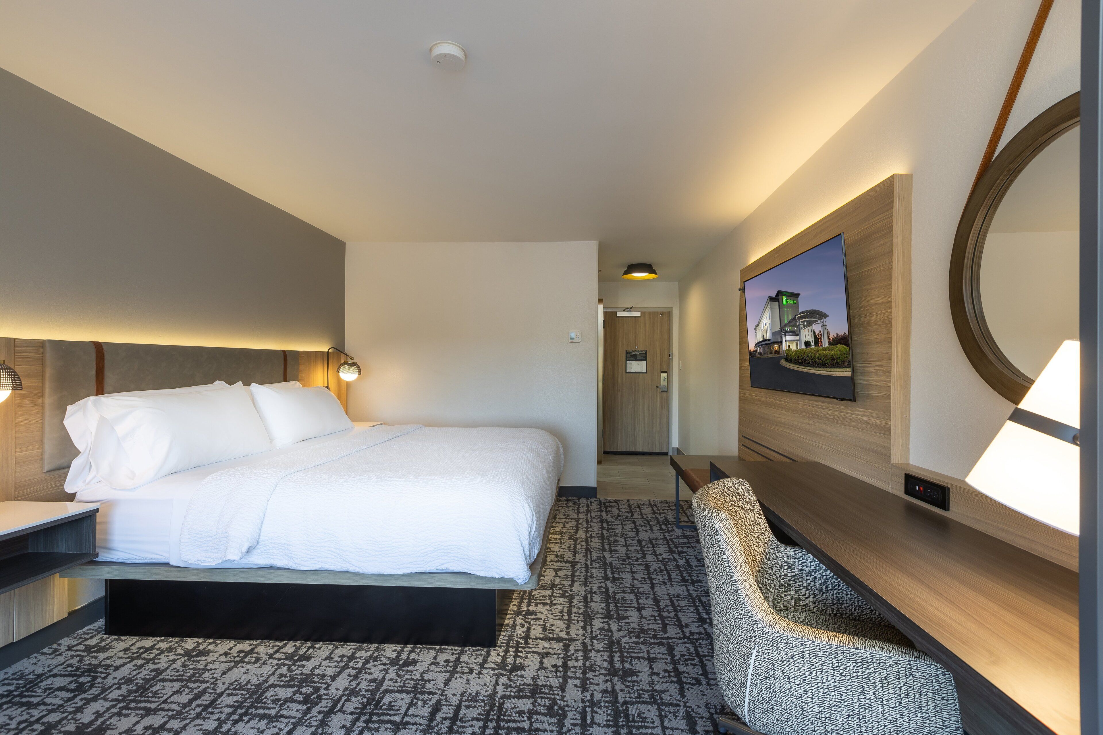 A hotel room with a large bed, bedside table, lamp, wall-mounted TV, and a mirror.