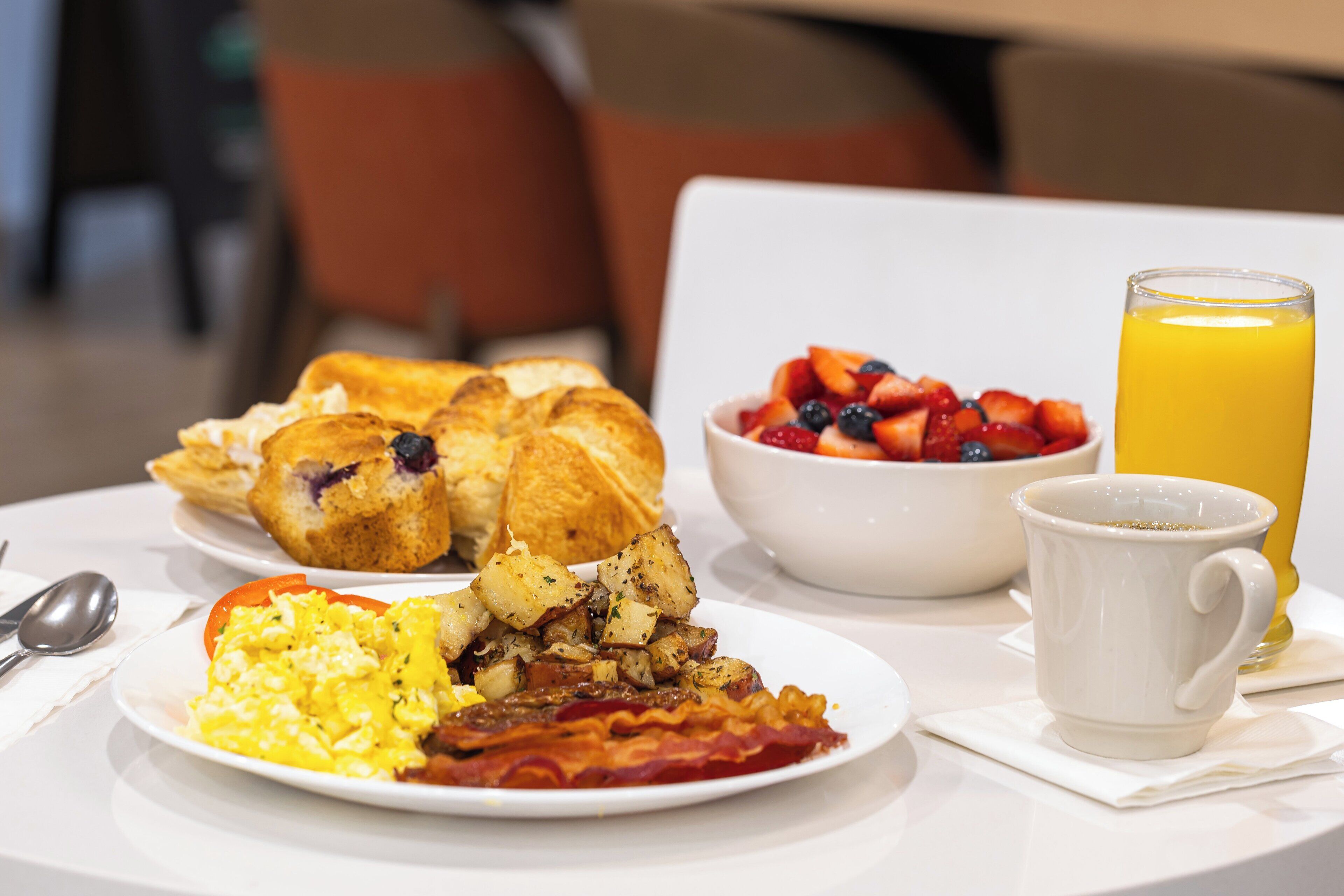 A breakfast table with plates of eggs, bacon, toast, and fruit, along with a glass of orange juice.