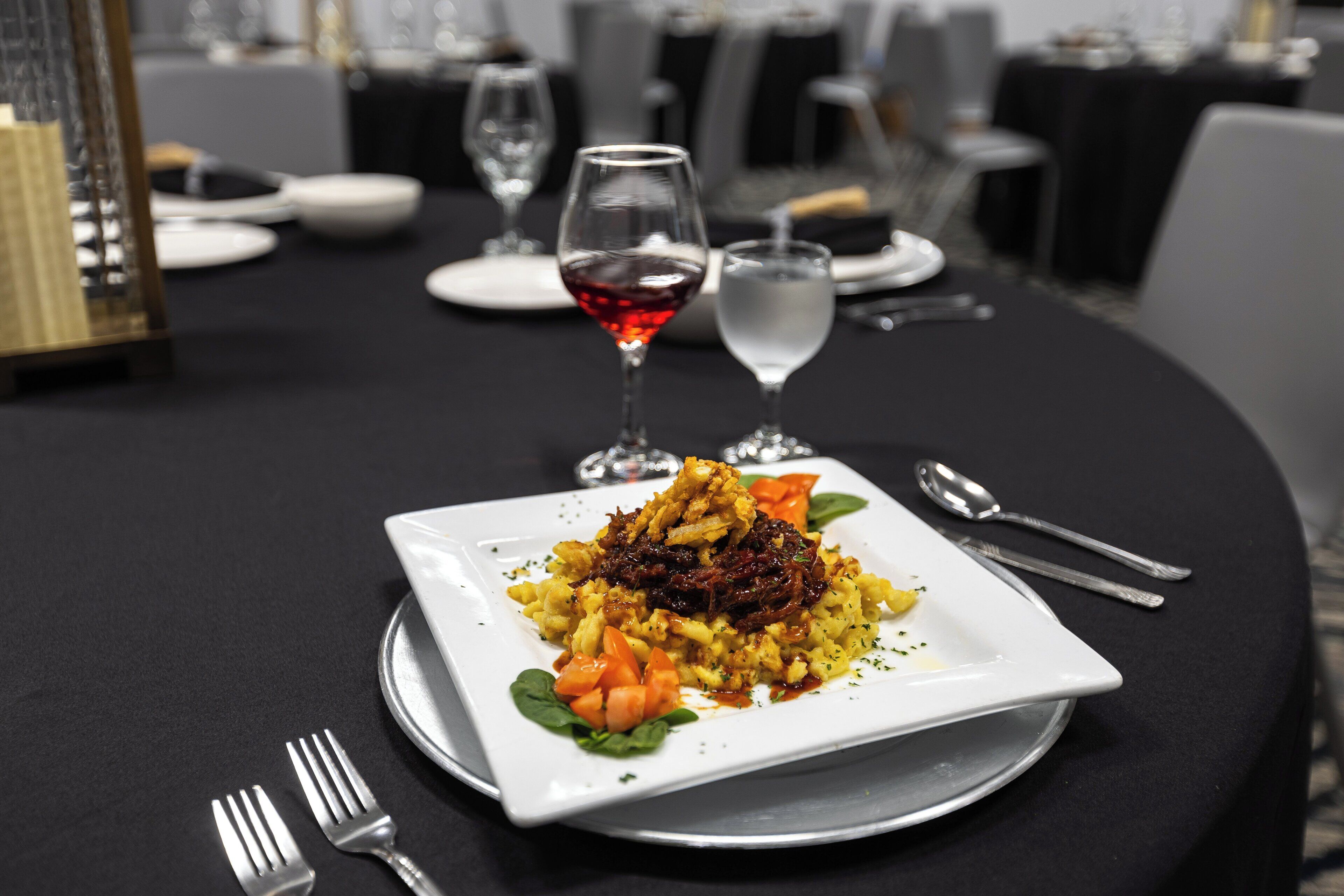 A square white plate with a dish of food, two forks, a wine glass with red wine, and a water glass on a table.