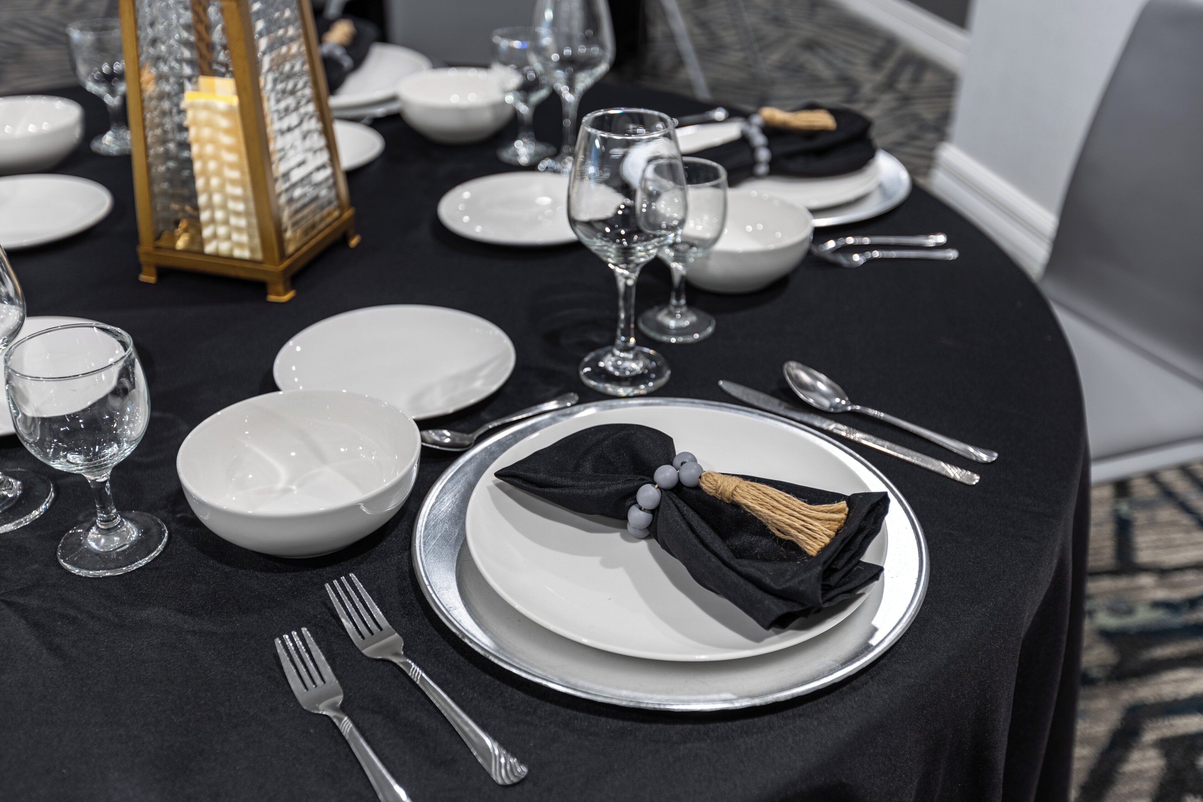 A table set with white plates, silverware, and clear glassware, with a black napkin and a decorative lantern in the background.