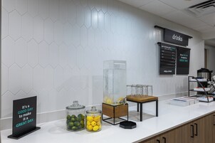 A breakfast bar with a sign, jars of lemons and limes, a juice dispenser, and a drinks menu.
