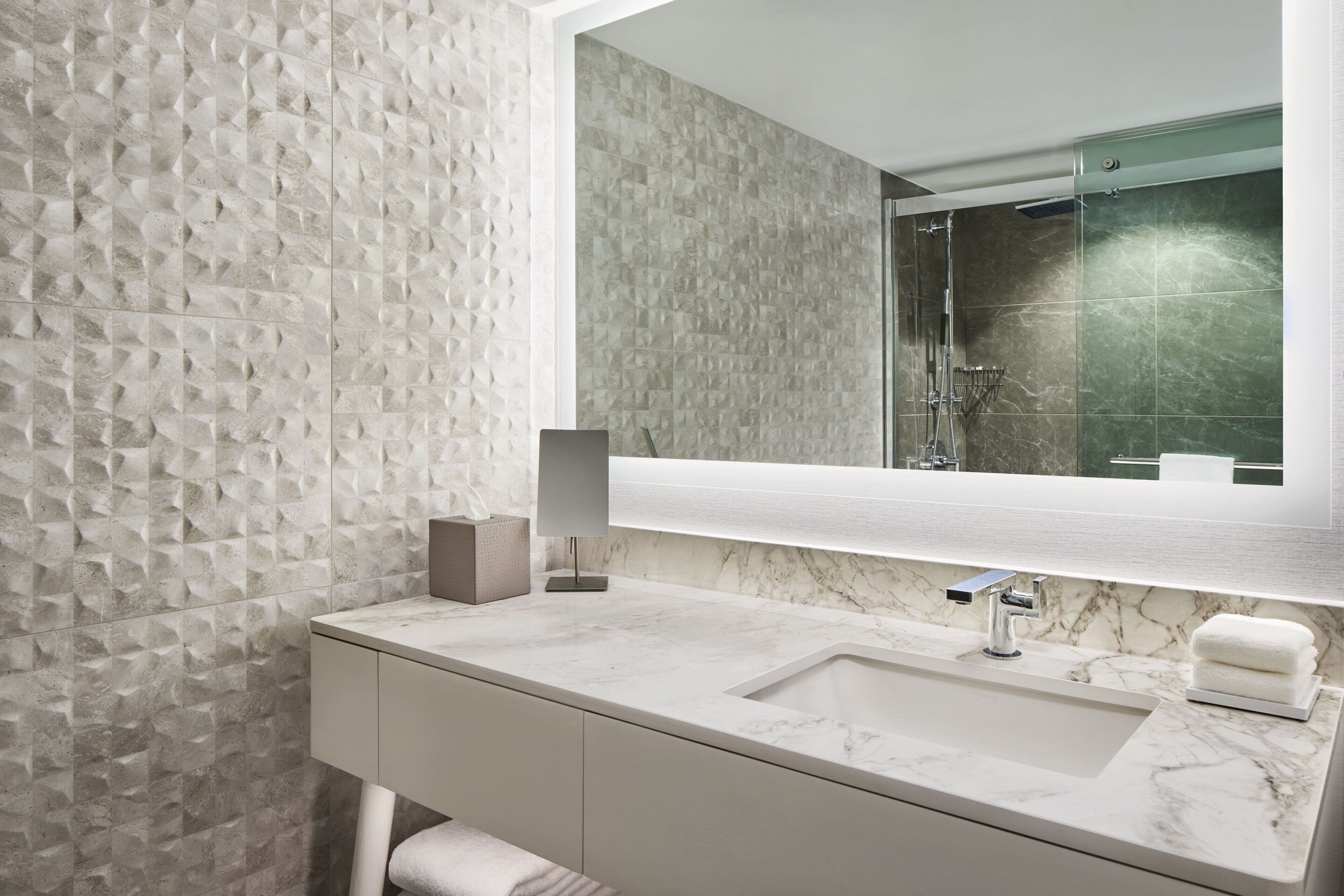 Un baño moderno con lavamanos de mármol, un espejo grande y azulejos con textura en la pared.