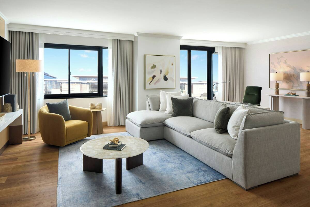 A modern hotel room with a large L-shaped sofa, a round coffee table, a yellow armchair, and a TV.