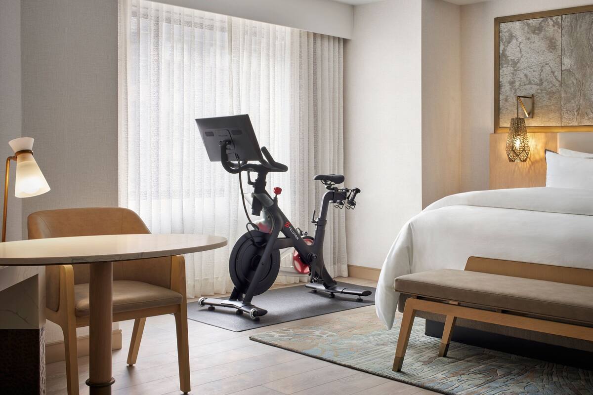 A hotel room with a bed, a desk, an exercise bike, and a chair.