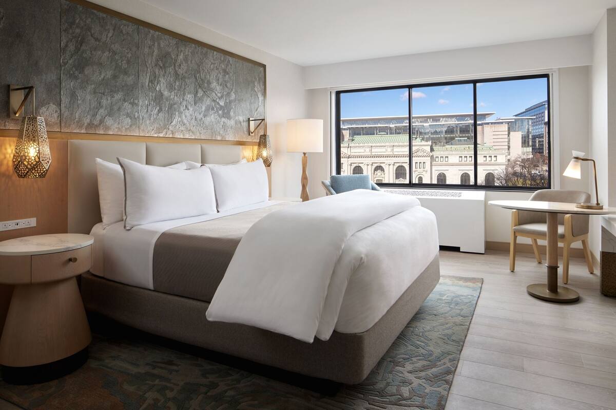 A hotel room with a large bed, a desk, a chair, a window with a city view, and a decorative wall panel.